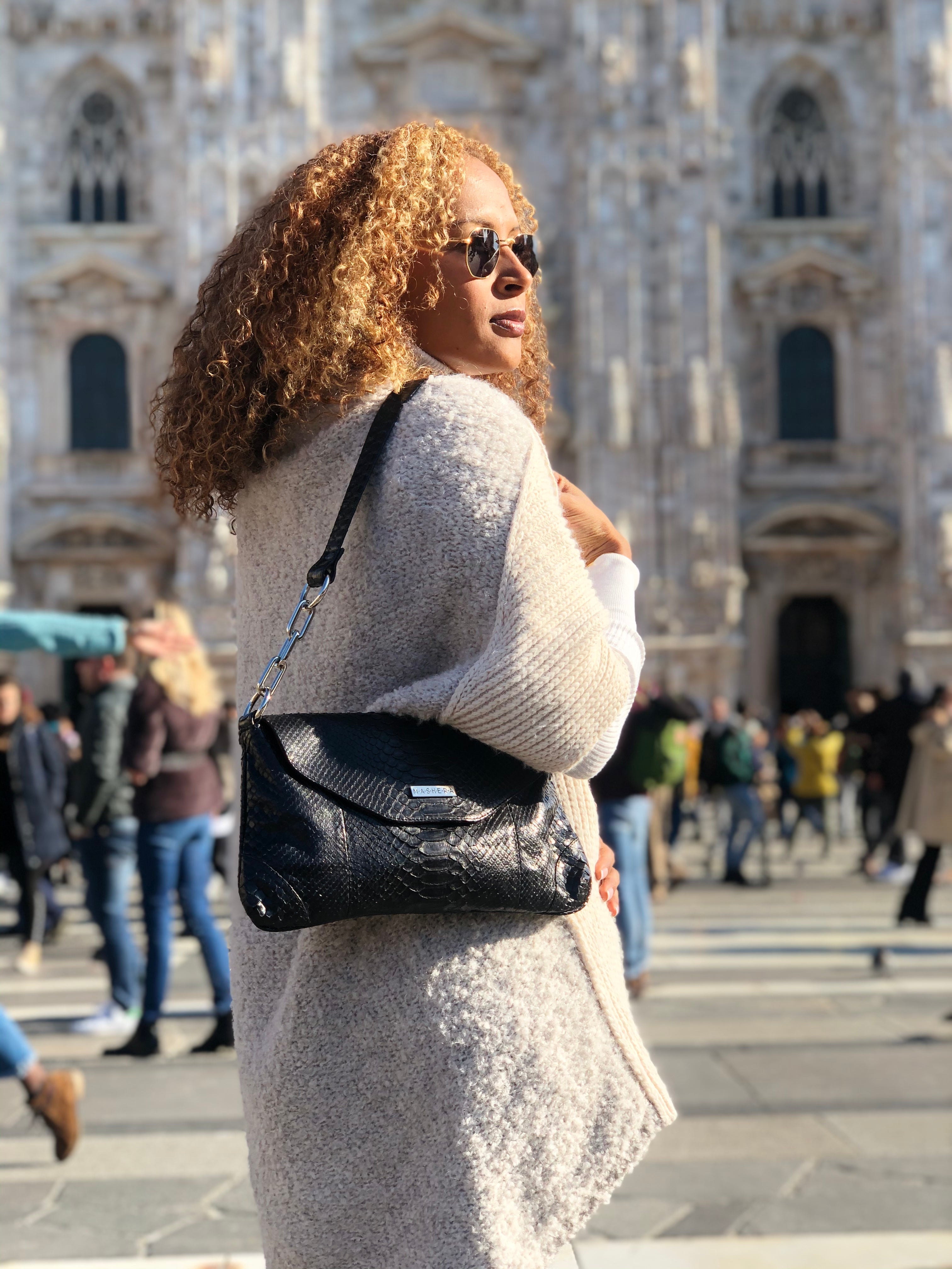 Modelo frente al Duomo de Milán luciendo el bolso CLEMENTINA negro en pitón - diseño emblemático con cadena y hombrera.